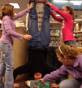 Mrs. Adair, Horizon librarian, reading a children's book while standing on her head, with the help of three students.