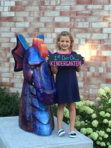Young female students stands next to a Dragon sculptur eon her first day of kindergarten.