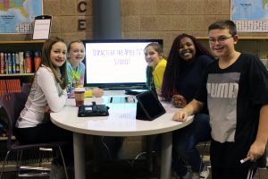 Five middle school students stand around a white board table smiling