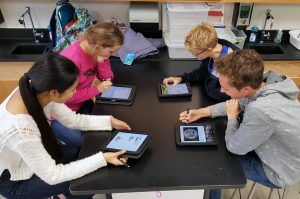 Four students sitting at a science table using ipads and talking.
