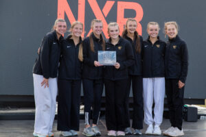 7 young women smile in front of black background with orange text holding plaque