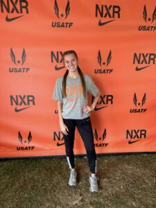 Girl smiles against orange background, wears gray shirt and black sweatpants